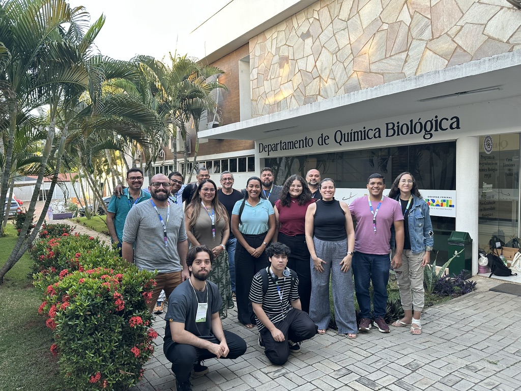 Grupo de 15 pessoas posa para foto em frente ao prédio do Departamento de Química Biológica, cercado por jardins com flores vermelhas e palmeiras. A fachada do prédio traz o nome do departamento em destaque, e os participantes sorriem, indicando um momento de confraternização, possivelmente relacionado a uma atividade acadêmica ou científica.