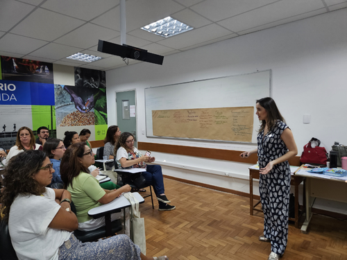 Foto em sala de aula. &Agrave; direita, uma mulher branca e de cabelos castanhos, vestida em um macac&atilde;o estampado, se direciona ao p&uacute;blico &agrave; esquerda, com diversas pessoas sentadas nas carteiras. Ao fundo, um quadro branco com um papel pardo.